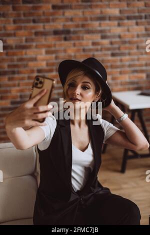 Elegante donna bionda con cappello nero sorridente fa selfie sul suo smartphone.Messa a fuoco selettiva morbida, sfocatura, rumore artistico. Foto Stock