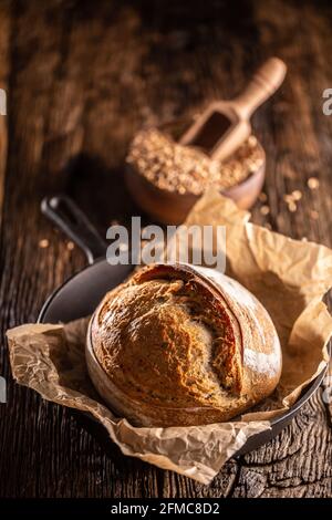 Pane rustico di lievito croccante appena sfornato sdraiato sulla cottura carta nel recipiente di legno in lega pieno di grano grani sessola di legno cucito profondamente in Foto Stock