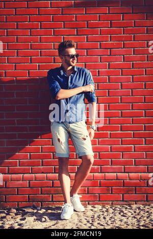 Giovane uomo che indossa una camicia blu e pantaloncini strappati un muro di mattoni Foto Stock