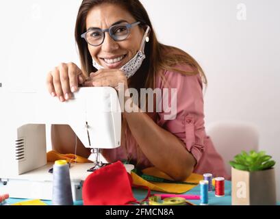 Donna felice che lavora con macchina da cucire facendo faccia protettiva casalinga maschera per prevenire e fermare il virus corona Foto Stock