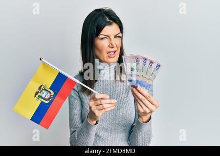 Giovane donna ispanica che detiene la bandiera della colombia e pesos colombiani banconote indecore e confuso espressione. Concetto di dubbio. Foto Stock