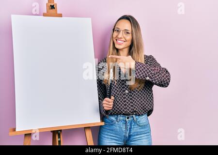 Bella donna ispanica in piedi da pittore cavalletto con spazzole sorridente, con mano e dito Foto Stock