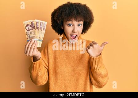 Giovane ragazza ispanica che tiene 5000 banconote forint ungheresi che puntano il pollice fino al lato sorridente felice con bocca aperta Foto Stock