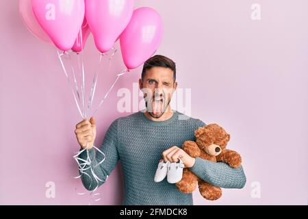 Bell'uomo con barba che si aspetta una bambina che tiene palloncini, scarpe e orsacchiotto che attaccano la lingua fuori felice con l'espressione divertente. Foto Stock