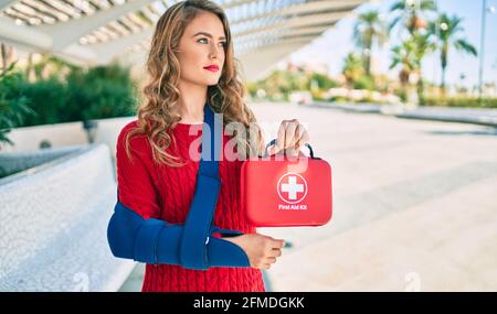 Giovane bionda con espressione seria infortunata con imbracatura a braccio e kit di pronto soccorso in mano al parco. Foto Stock