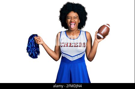 Giovane donna afroamericana che indossa uniforme cheerleader tenendo in mano il pompon e la palla di calcio che si attacca la lingua fuori felice con l'espressione divertente. Foto Stock