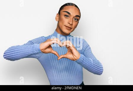 Uomo ispanico transgender che indossa make up e capelli lunghi indossando abiti casual sorridente in amore mostrando il simbolo del cuore e la forma con le mani Foto Stock