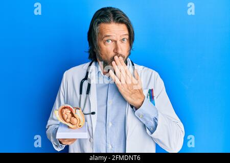 Bell'uomo ginecologo di mezza età che tiene modello anatomico dell'utero femminile con il feto che copre la bocca con la mano, scioccato e impaurito per errore. Su Foto Stock