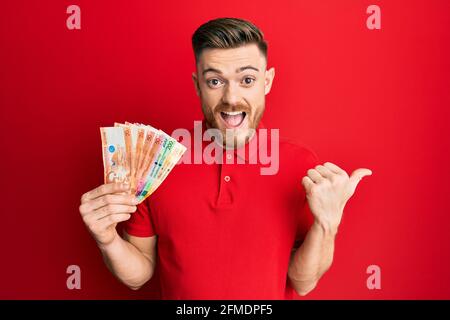 Giovane uomo rosso che tiene le banconote del peso filippino che punta il pollice in su al lato sorridente felice con bocca aperta Foto Stock