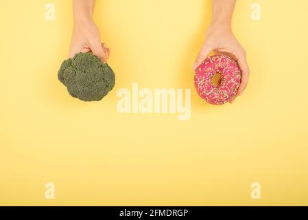 Confronto delle abitudini alimentari. Donna che tiene broccoli e ciambella su sfondo giallo. Spazio di cimasa. Foto Stock