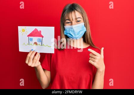 Adolescente caucasica ragazza che indossa maschera medica e tenendo casa disegnare sorridente, con mano e dito Foto Stock