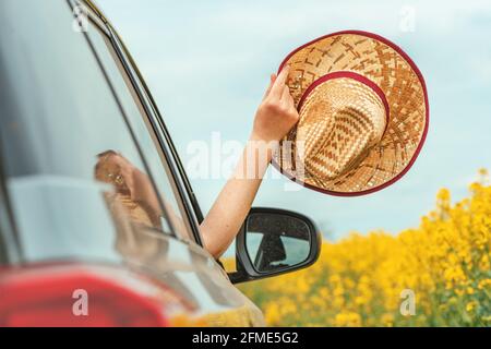 Donna che ama guidare in auto in un paesaggio fiorente della campagna estiva, mano con cappello di paglia che raggiunge il finestrino, fuoco selettivo Foto Stock