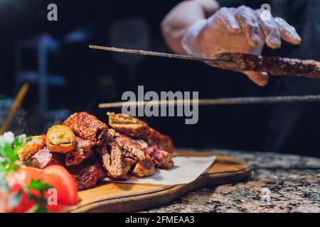Uomo in guanti neri mettere agnello con spatola per cibo su piatto bianco. Gustosa carne di agnello marrone cotta Foto Stock