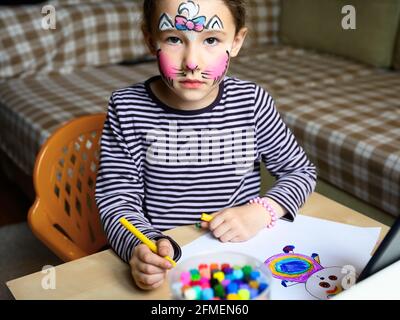 Disegno di capretto con le penne di feltro-punta interna, bambina con la maschera verniciata sul viso che studia a casa. Carino bambino impara a disegnare al tavolo in camera. Ritratto di Foto Stock