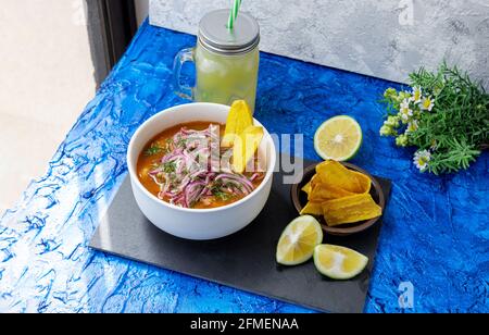 Piatto di encobollado, cibo tipico dell'Ecuador preparato con pesce, cipolla, limone e yucca su sfondo blu accanto ad una finestra con luce mattutina. Foto Stock