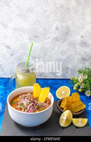 Piatto di encobollado, un alimento tipico dell'Ecuador preparato con pesce, cipolla e yucca con una caraffa di limonata e scaglie di banana su un blu e una ruggine Foto Stock