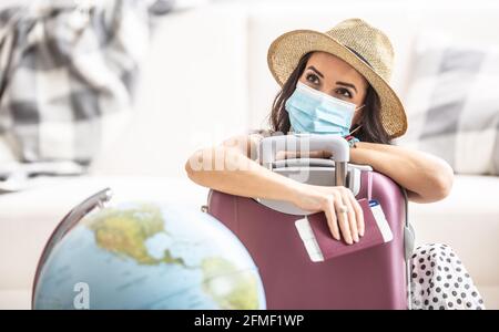 Donna in estate cappello e maschera viso si appoggia contro la valigia con biglietto di volo e un passaporto che pensa di viaggiare durante la pandemia. Foto Stock