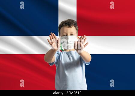 Piccolo ragazzo bianco in una maschera protettiva sullo sfondo della bandiera della Repubblica Dominicana fa un segno di stop con le mani, rimanere a casa Rep Domenicano Foto Stock