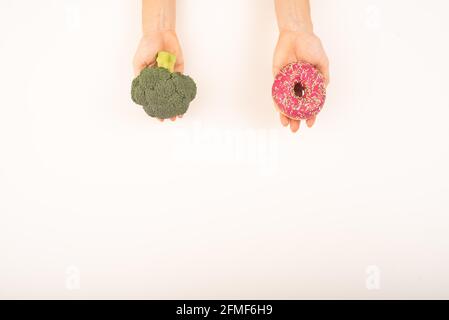 Confronto delle abitudini alimentari. Donna che tiene broccoli e ciambella su sfondo bianco. Spazio di copia Foto Stock
