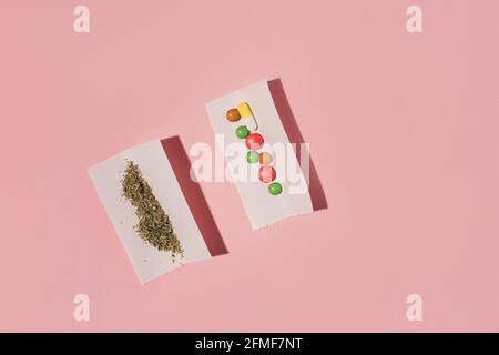 Due rotoli di carta con Marijuana secca e pillole colorate, piatto su sfondo rosa. Foto Stock