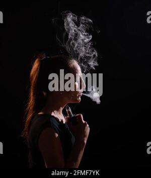 La bruna fuma una sigaretta elettronica su uno sfondo nero, rilascia fumo spesso dalla bocca. Ritratto di una donna che fuma un vape, che si abbonda. Foto Stock