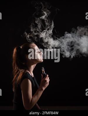 La bruna fuma una sigaretta elettronica su uno sfondo nero, rilascia fumo spesso dalla bocca. Ritratto di una donna che fuma un vape, che si abbonda. Foto Stock