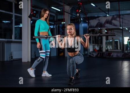 La giovane ragazza atletica si affolla con un barbell in palestra sotto la supervisione di un allenatore. L'allenatore femminile insegna affondo con bar. Personale Foto Stock