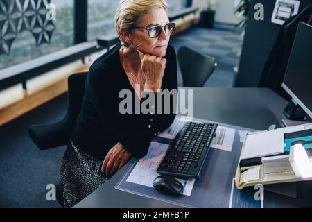 Donna d'affari anziana seduto alla sua scrivania guardando il computer. Donna con la mano sul mento al suo posto di lavoro. Foto Stock