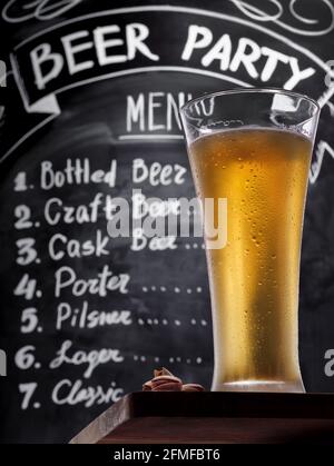 Bicchiere pieno di birra fredda e leggera con pistacchi su un tavolo di legno sullo sfondo di una tavola di gesso con testo del menu, vista dal basso verso l'alto. Birra alla spina Foto Stock