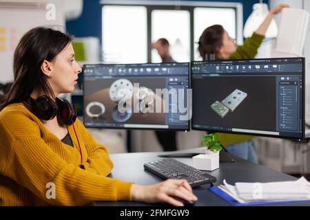 Ingegnere femminile industriale che guarda al personal computer con configurazione a doppio monitor, schermi che mostrano il software CAD con prototipo 3D di ingranaggi metallo pezzo meccanico. Dipendente che lavora in ufficio creativo Foto Stock