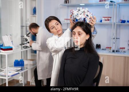 Tecnico di laboratorio medico che mette le cuffie eeg che monitorano le funzioni cerebrali, descovering la diagnosi di malattia. Gruppo di ricercatori che lavorano fino a tarda notte in clinica di neurologia per la salute della donna. Foto Stock