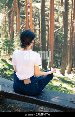 Giovane donna seduta su una panchina mentre medita in una foresta di alberi alti. Verticale. Foto Stock