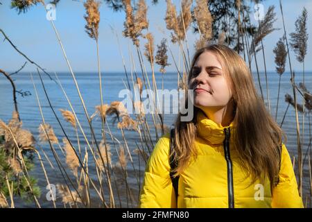 Portret di felice sorridente giovane donna con gli occhi chiusi gode del sole e aria fresca. Si leva in piedi contro il lago il caldo giorno di primavera in giallo Foto Stock