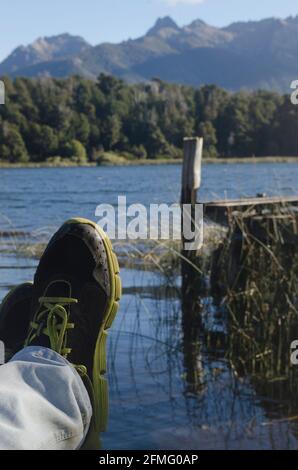 Gambe con jean e pantofole poggiate sulla riva del lago con un mullo accanto, montagna e laghi Patagoni vicino alla città di Bariloche Foto Stock