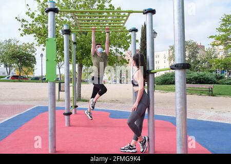 Due amici oscillano in una barra delle scimmie in un parco calistenico indossando maschere facciali protettive. Fitness all'aperto. Foto Stock