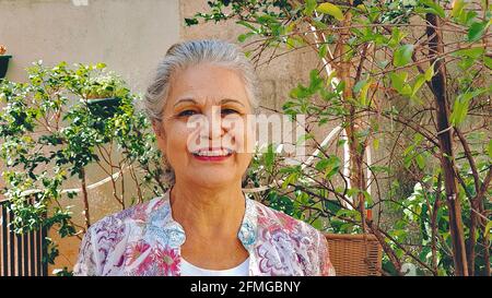 Donna matura che sorride nella casa del cortile con sole e piante. Donna bianca con capelli grigi a casa. Foto Stock