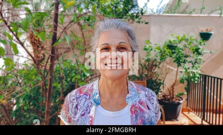 Donna matura che sorride nella casa del cortile con sole e piante. Donna bianca con capelli grigi a casa. Foto Stock