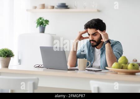 Ha sottolineato l'imprenditore arabo maschio che ha problemi al lavoro, seduto turbato con il computer portatile al tavolo da cucina Foto Stock