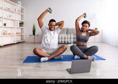 Felice addestramento della coppia nera con i manubri vicino al laptop a casa, facendo il fitness in linea al video di sport Foto Stock