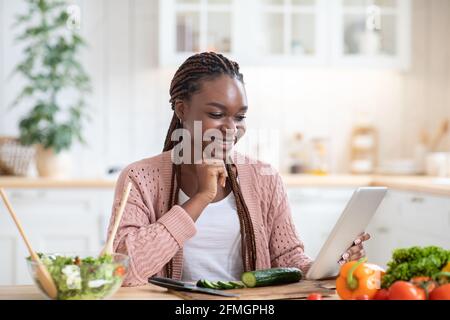 Buona donna africana che controlla la ricetta su tablet digitale durante la cottura in cucina Foto Stock