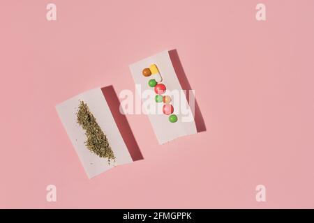 Due rotoli di carta con Marijuana secca e pillole colorate, piatto su sfondo rosa. Foto Stock