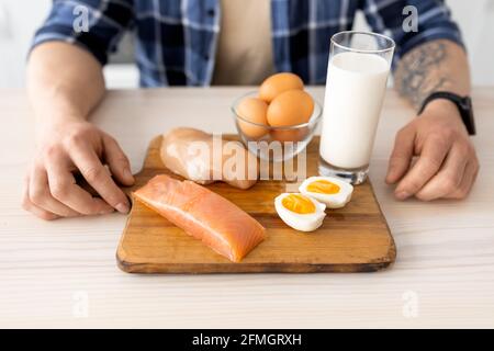 Mangiare sano. Uomo che prepara la colazione, seduto a tavola con filetto di carne crudo, pesce, latte e uova su tavola di legno Foto Stock