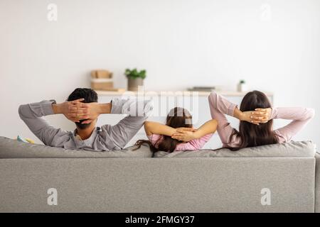 Tempo libero. Vista posteriore di genitori rilassati e bambino con le braccia dietro la testa seduta sul divano e riposante a casa Foto Stock