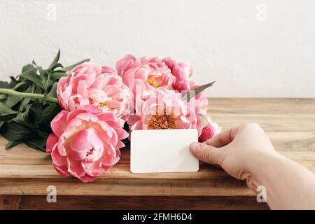Mano della donna che tiene in bianco saluto, biglietto da visita. Fiori di peonie rosa, bouquet sdraiato su vecchio tavolo di legno. Invito mock-up. Parete bianca. Matrimonio o. Foto Stock