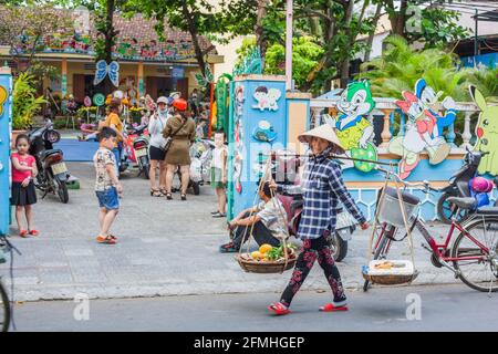 Femmina vietnamita che porta due cestini sulla spalla palo oltre la scuola dei bambini nella città vecchia, Hoi An, Vietnam, Foto Stock