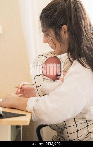 Vista laterale della madre con il bambino piangente nel bambino portatore seduto al tavolo in casa Foto Stock