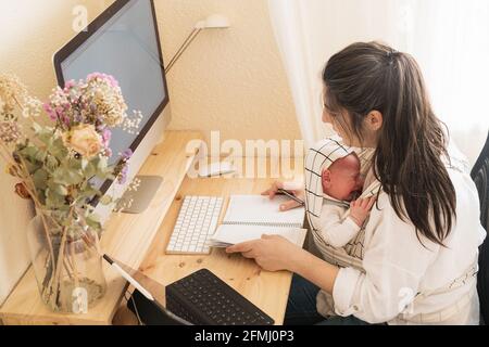 Madre adulta seduta alla scrivania che lavora su un computer desktop e. prendere appunti in notebook mentre si tiene piangendo bambino piccolo a. tavolo di giorno Foto Stock