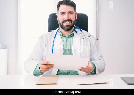 Medico maschio maturo amichevole con foglio di carta e stetoscopio aspetto alla macchina fotografica alla scrivania in clinica Foto Stock