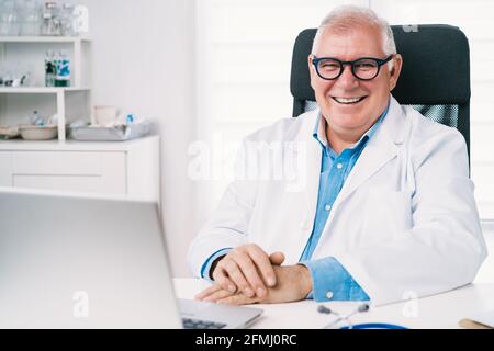 Felice sorridente anziano medico maschile in uniforme e eyewear tipizzazione su netbook al tavolo in clinica Foto Stock