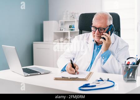 Medico maschile senior in uniforme che parla sul cellulare mentre prende appunti su carta alla scrivania con computer portatile in ospedale Foto Stock